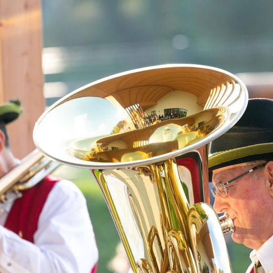 Ein älterer Mann mit Brille und Hut spielt ein Brass-Tuba. Ein anderer Mann in traditioneller Kleidung steht hinter ihm und spielt ebenfalls ein Blasinstrument.