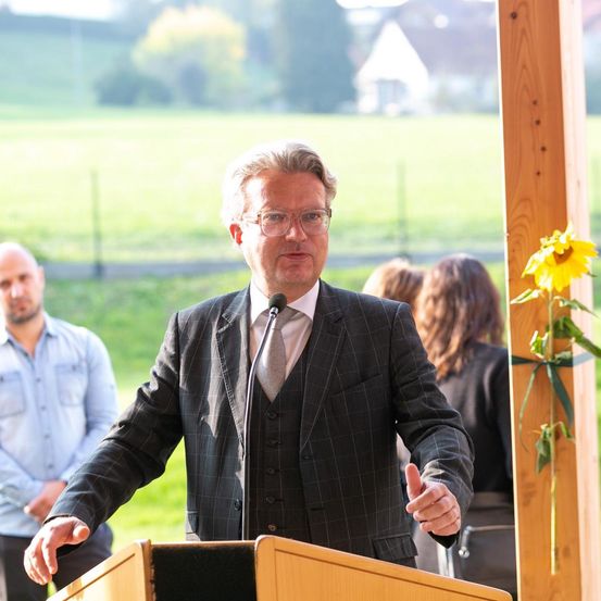 Ein Mann in Anzug und Brille steht an einem Podium und spricht in ein Mikrofon. Hinter ihm lauschen zwei Personen aufmerksam. Ein Sonnenblume schmückt das Podium.