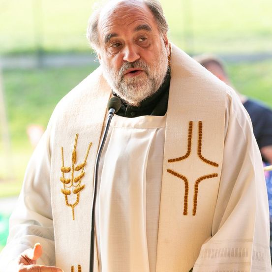 Ein Priester in einer weißen Robe mit einem goldenen Kreuz spricht in ein Mikrofon, im Freien mit einem verschwommenen grünen Feld und Menschen im Hintergrund.