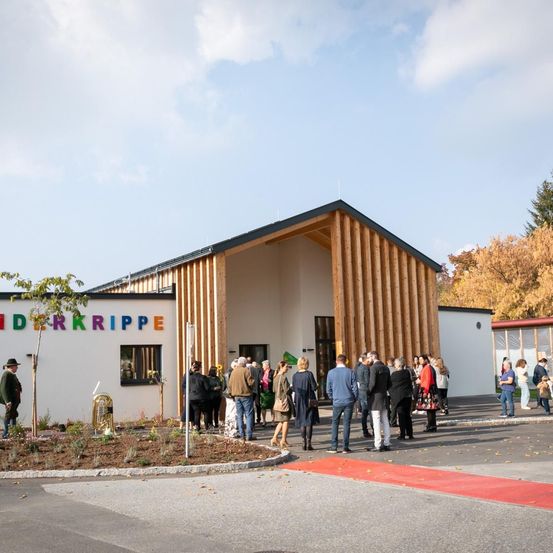 Eine Gruppe von Menschen steht vor einem Gebäude namens Kinderkrippe, umgeben von Bäumen, einem roten Teppich und einem Messinginstrument.