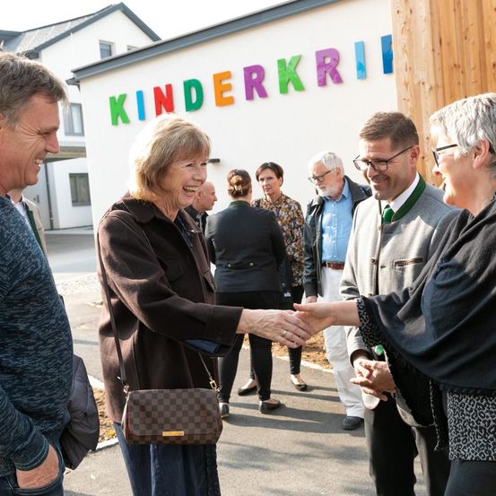 Eine Gruppe von Menschen steht vor einem Gebäude mit dem Wort Kinderkrippe. Eine Frau mit einer karierten Tasche gibt einem Mann die Hand.