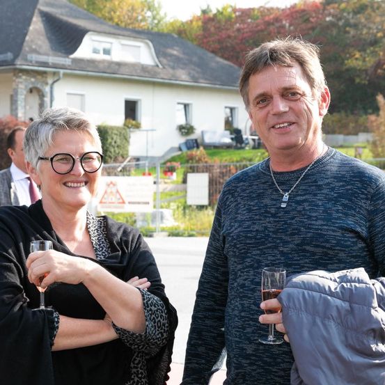 Ein Mann und eine Frau stehen lächelnd draußen mit Getränken in der Hand. Sie stehen vor einem Haus mit Garten, und im Hintergrund befinden sich Bäume und ein Zaun.