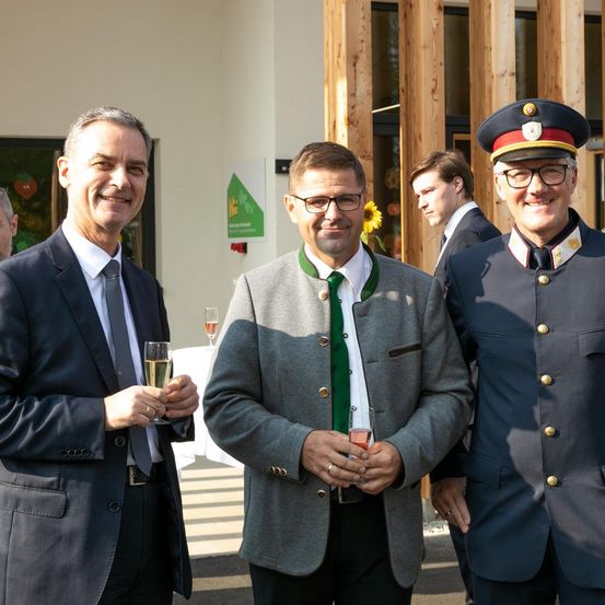 Drei Männer in formeller Kleidung stehen lächelnd vor einem Gebäude. Zwei von ihnen halten Gläser mit Wein, während der andere ein Glas mit roter Flüssigkeit hält. Hinter ihnen stehen weitere Personen, und eine Blume ist sichtbar.