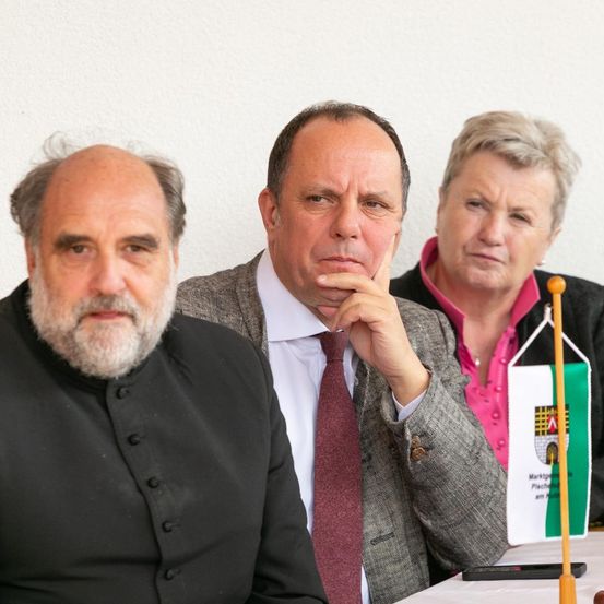 Drei Personen sitzen an einem Tisch, einer mit Bart, einer mit der Hand am Kinn und eine Frau mit kurzen Haaren. Sie sind feierlich gekleidet.