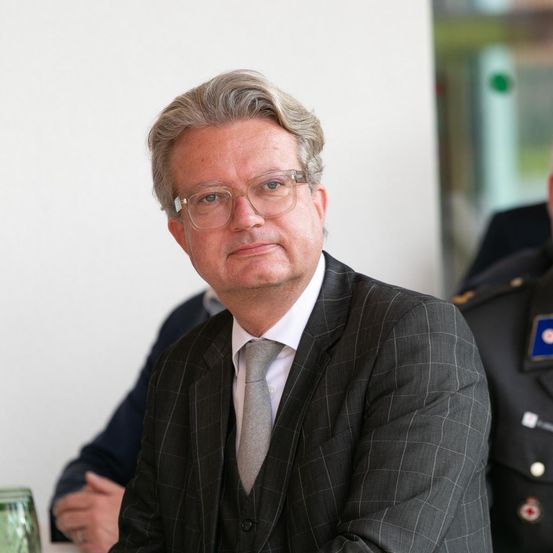 Ein Mann in Anzug und Brille sitzt aufmerksam bei einem Treffen, mit einem weiteren Mann in Uniform und einem verschwommenen Hintergrund.