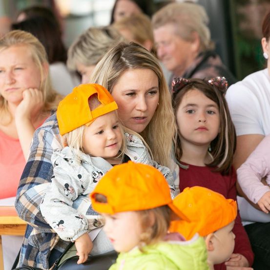 Eine Gruppe von Frauen und Kindern ist versammelt, einige tragen orangefarbene Mützen. Eine Frau hält ein Kind, das eine Mickey-Mouse-Jacke trägt. Andere Kinder sitzen neben ihnen, mit weiteren Menschen im Hintergrund.