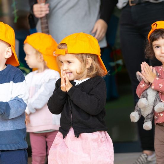 Eine Gruppe von Kindern mit orangefarbenen Mützen, die klatschen. Ein Mädchen in Schwarz mit einer rosa Hose lächelt. Dahinter hält eine Person eine Gitarre.