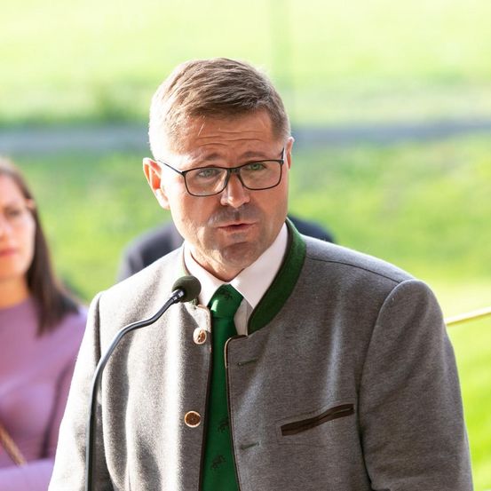 Ein Mann mit Brille und grüner Krawatte spricht in ein Mikrofon, er steht vor einem unscharfen Hintergrund mit grünem Gras und einer Person in einem lila Hemd.