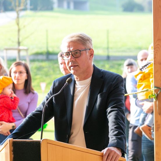 Ein Mann steht an einem Rednerpult mit einem Mikrofon. Er trägt eine Brille und einen dunklen Anzug. Hinter ihm stehen mehrere Menschen, darunter eine Frau mit einem Kind. Die Szene ist im Freien mit einem Rasen im Hintergrund.