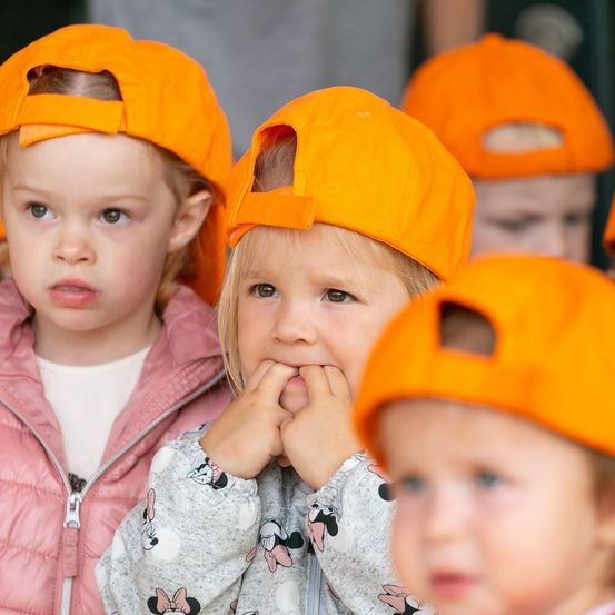 Eine Gruppe von Kindern, die orangefarbene Mützen tragen, möglicherweise bei einer Schulveranstaltung. Das Mädchen in der Mitte hat ein Minnie-Mouse-Motiv auf ihrer Jacke.