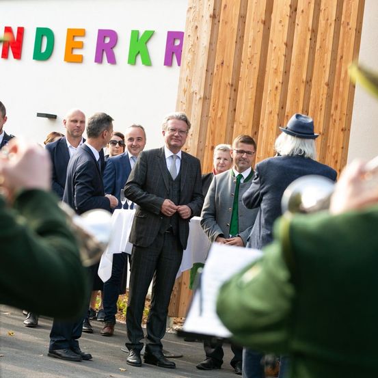 Eine Gruppe formell gekleideter Männer und Frauen steht vor einem Gebäude mit dem Wort Wunderkr an der Wand. Ein Mann hält ein Weinglas.