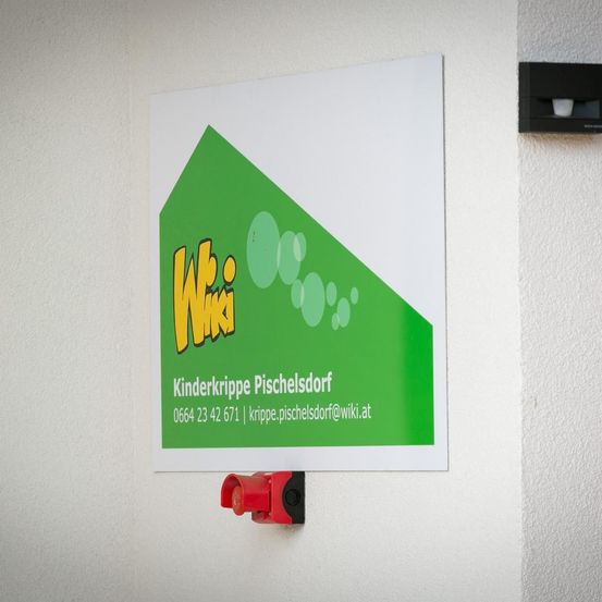 Ein Schild mit dem Text 'Wiki Kinderkrippe Pischelsdorf' und Kontaktinformationen ist an einer weißen Wand befestigt. Das Schild hat ein grünes Dreieck mit Blasen und einen roten Knopf darunter.