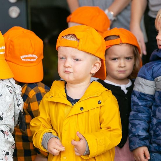 Eine Gruppe junger Kinder in orangefarbenen Mützen und gelben Regenmänteln steht zusammen, wobei ein Kind ernst aussieht.