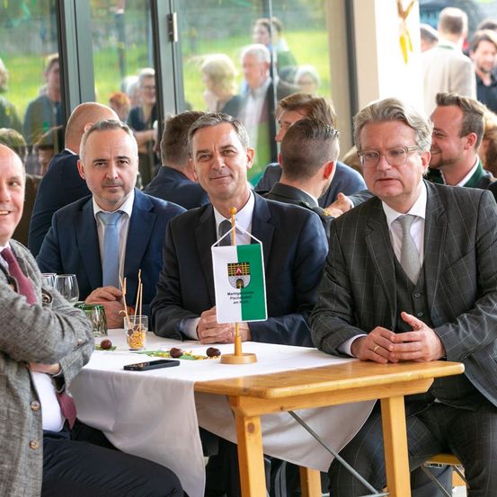 Vier Männer in Anzügen sitzen an einem Tisch, wahrscheinlich bei einer Konferenz. Einer hält eine Flagge und ein anderer hat die Hände gefaltet. Hinter ihnen stehen und sitzen Menschen in einem Raum mit Glaswänden.