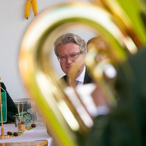 Ein Mann mit Brille sitzt an einem Tisch, mit einer goldenen Tuba im Vordergrund. Hinter ihm ist eine weitere Person teilweise sichtbar. Mais hängt an der Wand.