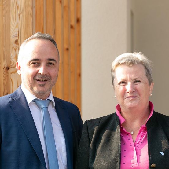 Ein Mann und eine Frau stehen nebeneinander und lächeln in die Kamera. Der Mann trägt einen dunkelblauen Anzug mit einer hellblauen Krawatte. Die Frau trägt ein rosa Hemd und eine schwarze Jacke mit einem Anstecker auf der linken Seite. Hinter ihnen ist eine Holzwand.