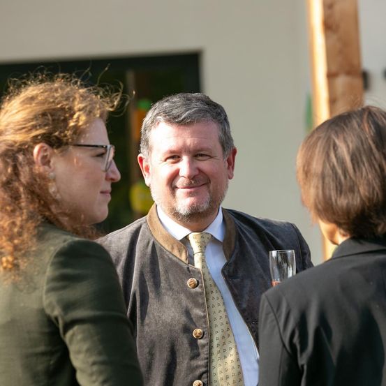 Ein Mann mit lächelndem Gesicht steht zwischen zwei Frauen vor einem Gebäude, eine hält ein Glas.