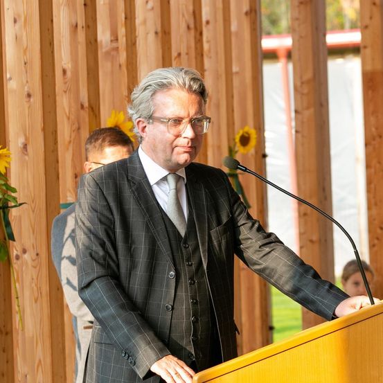 Ein Mann in formeller Kleidung steht an einem Rednerpult mit Mikrofon und spricht eine Zuhörerschaft an. Hinter ihm steht eine weitere Person in der Nähe einer Holzwand, die mit Sonnenblumen geschmückt ist.