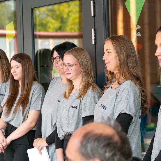 Fünf Frauen in gleichen grauen T-Shirts stehen vor einem Gebäude und schauen nach vorne. Zwei Frauen tragen Brillen. Das Gebäude hat Glastüren und ein farbenfrohes Drachendesign.