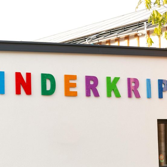 Ein farbenfrohes Wort 'Kinderkrippe' auf einer weißen Wand, mit Buchstaben in verschiedenen hellen Farben. Die Wand befindet sich in der Nähe eines Gebäudes mit einem Metalldach.
