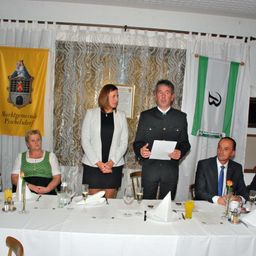 Eine formelle Versammlung mit vier Personen, die an einem Tisch sitzen. Ein Mann steht und hält ein Papier. Zwei Flaggen hängen hinter ihnen.