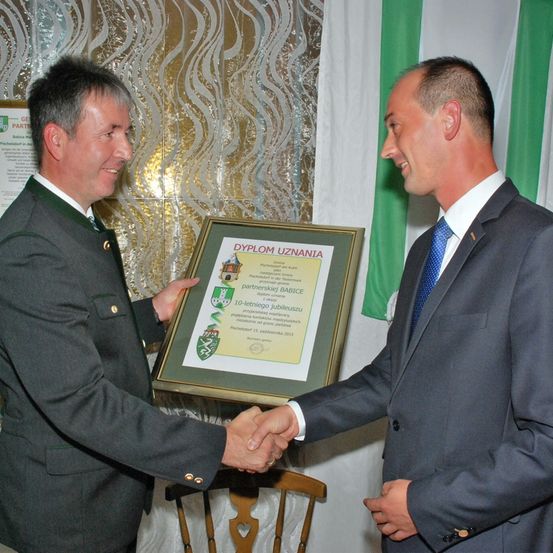 Zwei Männer, einer in Uniform und der andere in Anzug, schütteln sich die Hände, während sie ein gerahmtes Zertifikat vor einem grün-weißen Hintergrund halten.