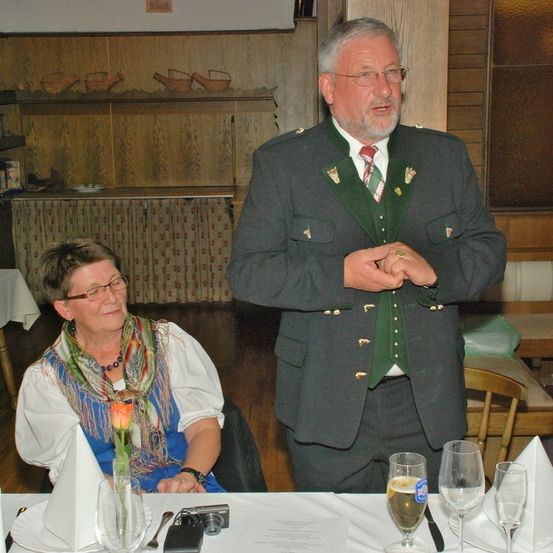 Ein Mann in traditioneller deutscher Uniform steht und spricht an einem Esstisch mit einer sitzenden Frau, einer Blumenvase und Weingläsern.