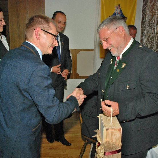 Zwei Männer schütteln sich die Hände, während ein anderer Mann eine Holzkiste hält, drinnen. Hinter ihnen unterhält sich ein Mann mit einer anderen Person. Ein Banner mit einem Bild ist an der Wand.