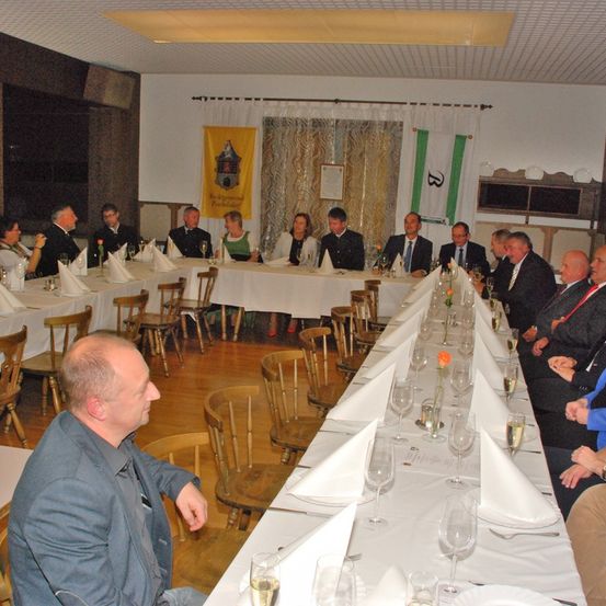Eine Gruppe von Menschen in Anzügen und Krawatten sitzt an einem langen Tisch mit Weinglas und Servietten. Einige tragen Krawatten, andere Brillen. Ein Banner hängt hinter ihnen.