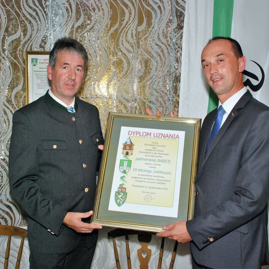 Zwei Männer in Uniformen stehen und halten ein Zertifikat. Der Mann auf der linken Seite trägt eine Medaille, während der Mann auf der rechten Seite einen Anzug trägt. Dahinter befindet sich ein Hintergrund mit einer Flagge und einem gerahmten Zertifikat.