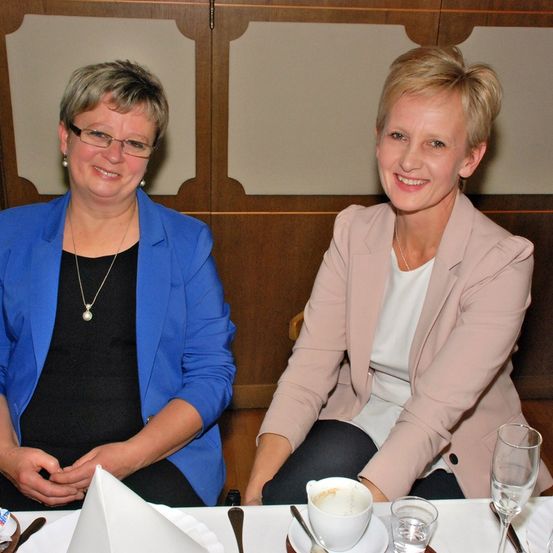 Zwei Frauen sitzen in einem Restaurant an einem Tisch und lächeln für ein Foto. Beide tragen helle Blazer. Die linke trägt einen blauen Blazer, die rechte einen rosa Blazer. Auf dem Tisch stehen Tassen, Untertassen und Gläser.
