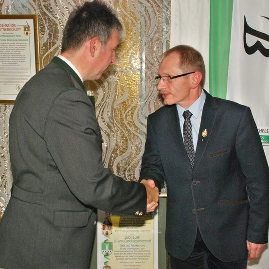 Zwei Männer schütteln sich die Hände vor einer Wand mit einem gerahmten Dokument. Ein Mann trägt eine grüne Weste.