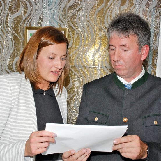 Eine Frau und ein Mann sehen sich ein Stück Papier an. Die Frau hat kurze braune Haare und trägt eine weiße Jacke. Der Mann trägt einen grauen Anzug und hat graues Haar. Hinter ihnen ist eine goldene Wand mit Muster.