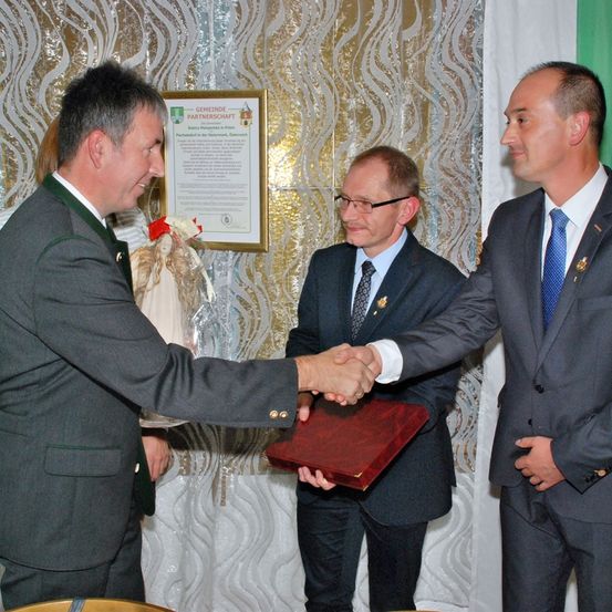 Drei Männer schütteln sich die Hände. Der Mann auf der linken Seite hält ein Geschenk. Der Mann in der Mitte hält eine rote Mappe. Sie stehen vor einer Wand mit einem gerahmten Zertifikat.