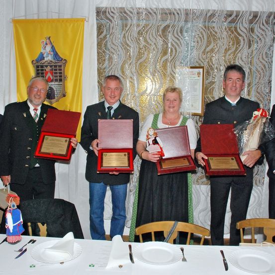 Fünf Menschen in formeller Kleidung stehen vor einem Tisch und halten Plaketten in roten Schachteln. Sie lächeln und scheinen ein Ereignis zu feiern. Hinter ihnen ist ein gelber Banner mit einem Logo.