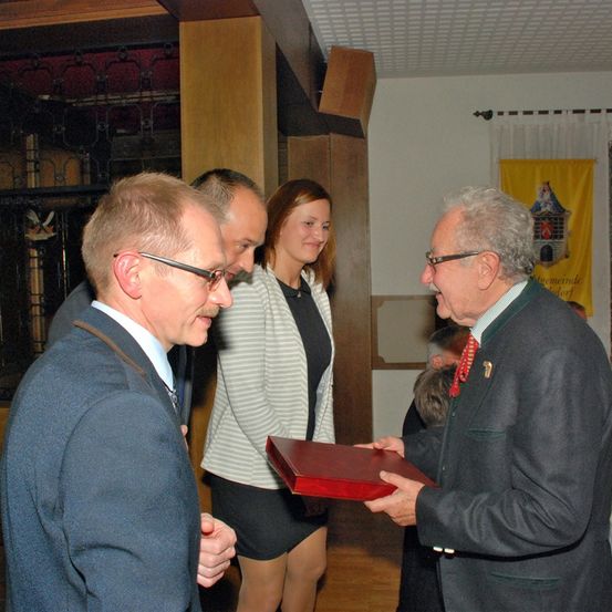 Ein Mann überreicht einem anderen Mann einen roten Ordner in einer formellen Umgebung. Eine Frau steht zwischen ihnen.