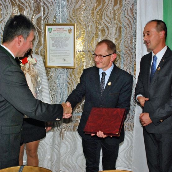 Drei Männer in Anzügen schütteln sich die Hände. Einer hält ein rotes Buch. Hinter ihnen befindet sich ein gerahmtes Zertifikat und ein weißer Vorhang.
