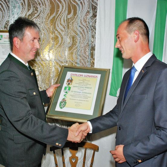Zwei Männer in Anzügen, einer mit einer Namensplakette, halten ein gerahmtes Zertifikat und schütteln sich die Hände vor einem grün-weißen Hintergrund.