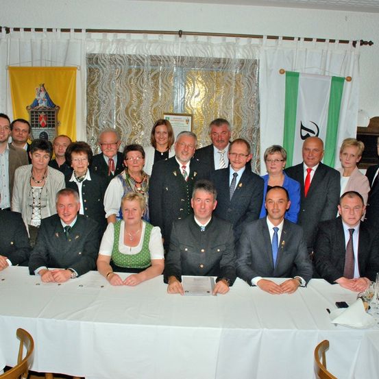 Eine Gruppe von Menschen in formeller Kleidung, sitzend und stehend um einen Tisch mit weißer Tischdecke, wahrscheinlich bei einer formellen Veranstaltung.