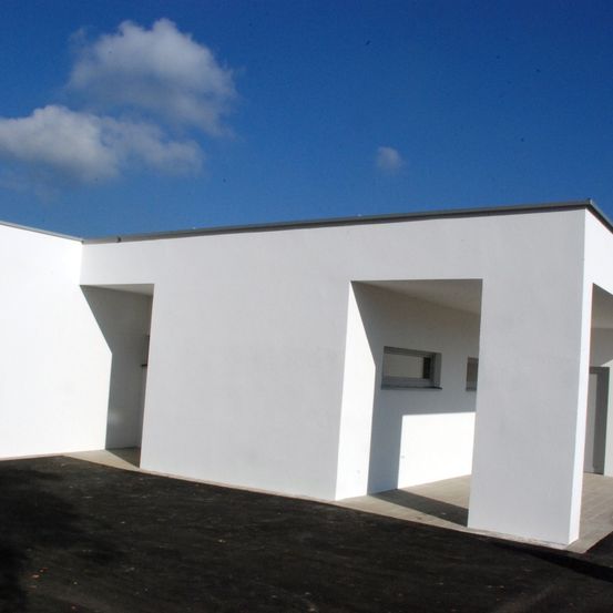 Ein weißes modernes Gebäude mit zwei Eingängen und einem Fenster, unter einem blauen Himmel mit einigen Wolken.