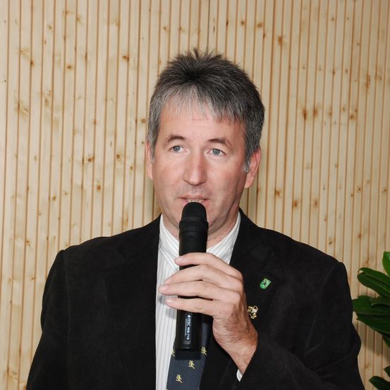 Ein Mann mit grau werdendem Haar und einem Schnurrbart spricht in ein Mikrofon. Er trägt einen schwarzen Anzug, ein weißes Hemd und eine Krawatte mit einer grünen Anstecknadel. Die hölzerne Wand hinter ihm hat eine strukturierte Oberfläche.