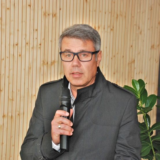 Ein Mann mit Brille und schwarzem Anzug hält ein Mikrofon und scheint zu sprechen. Hinter ihm befindet sich eine Holzwand und eine Pflanze.