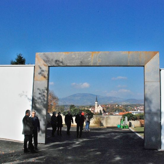 Eine Gruppe von Menschen geht durch einen Betonbogen, umgeben von blauem Himmel und Grün, mit Bergen in der Ferne.