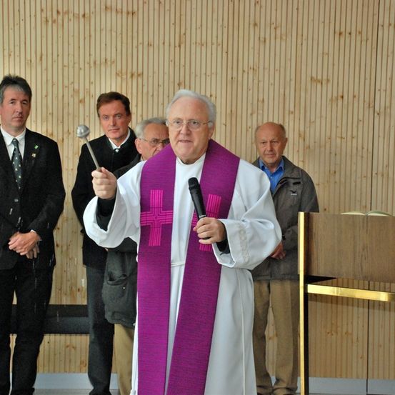 Ein älterer Priester in einer weißen Robe und einem violetten Schal spricht in ein Mikrofon, während eine Gruppe von Männern in formeller Kleidung hinter ihm steht.