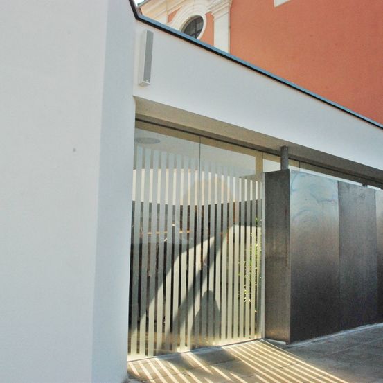 Das Bild zeigt ein modernes architektonisches Design mit einer weißen Wand und einer Glastür. Ein Metalltor ist teilweise geöffnet, und Sonnenlicht strömt durch und wirft Schatten.