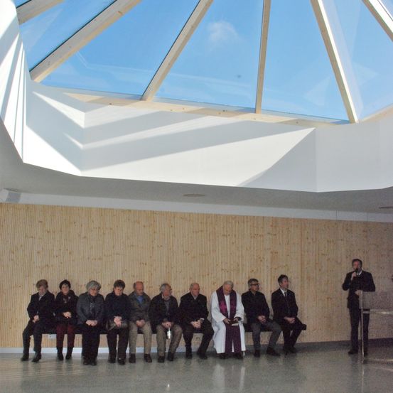 Eine Gruppe von Menschen sitzt auf Bänken in einem Raum mit einer Holzwand und einer Glasdecke. Ein Priester steht vor ihnen.