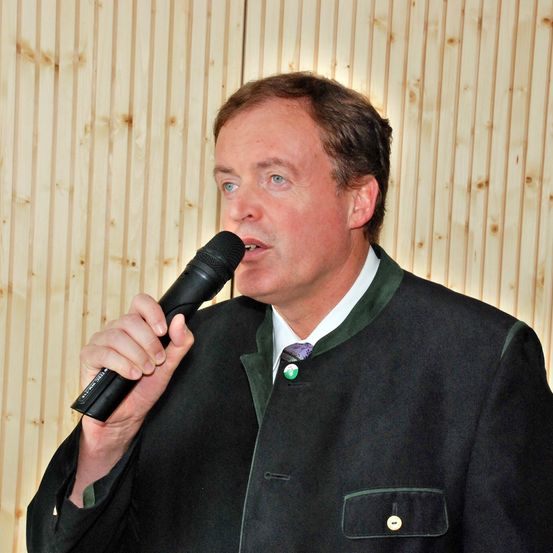 Ein Mann in einem dunklen Anzug und grüner Weste spricht in ein Mikrofon, vor einer Holzwand stehend.