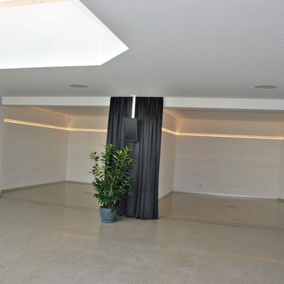 Ein leerer Raum mit weißen Wänden und Böden hat eine Decke mit einem Oberlicht. Ein hoher Topf steht in der Nähe schwarzer Vorhänge auf der rechten Seite.