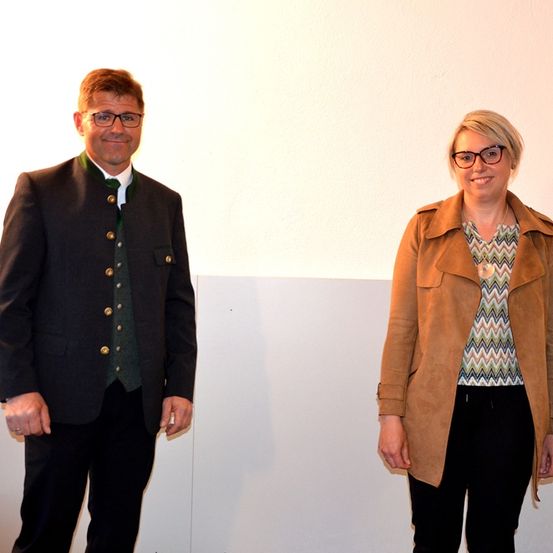 Ein Mann und eine Frau stehen nebeneinander, beide lächelnd, vor einer schlichten weißen Wand.