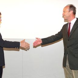 Zwei Männer in Anzügen und Krawatten, einer mit Brille, lächeln und schütteln sich in einer formellen Umgebung die Hände.